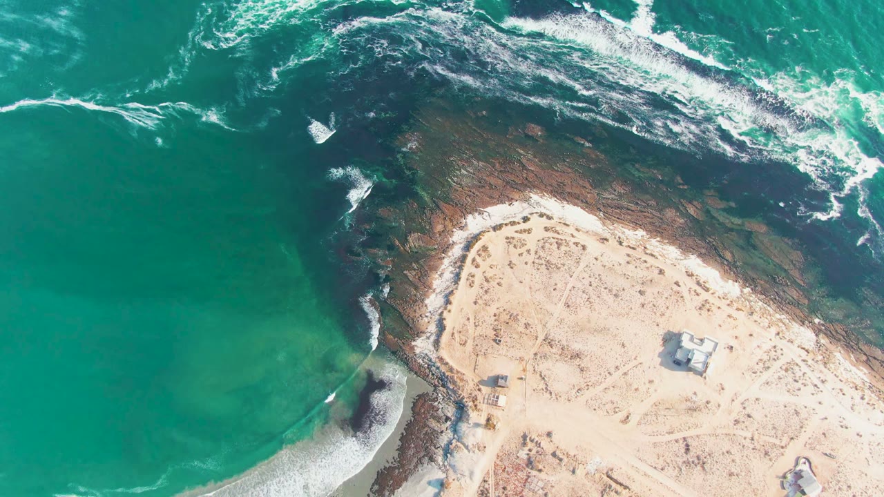 Top-down aerial frame of the La Bocana shoreline with turquoise water on the left, foamy wave streaks above the reef, pale sand on the right, and no visible people.