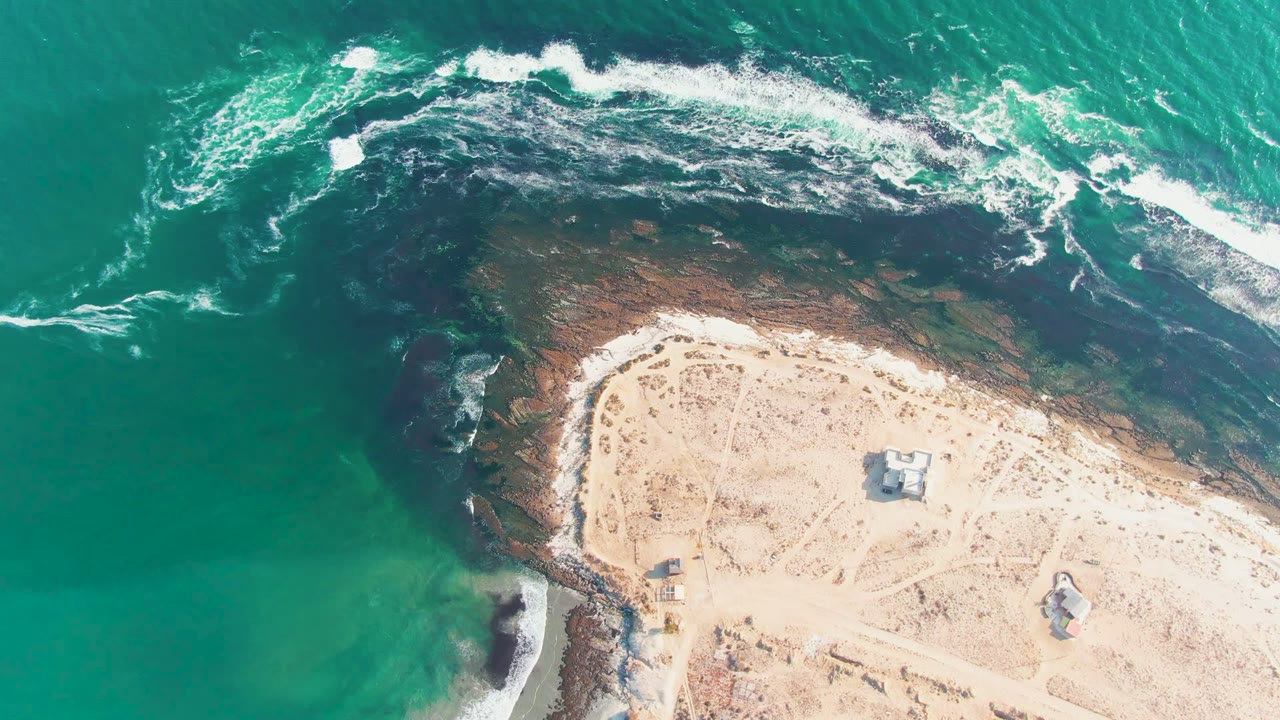 Overhead frame from La Bocana, Baja California Sur showing turquoise water and white surf curling around dark rocks beside pale sand, with no visible people.