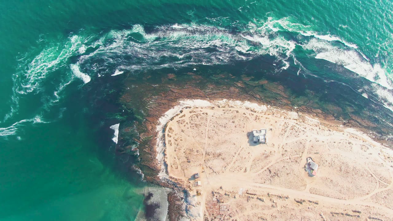 Aerial frame over La Bocana, Baja California Sur, with turquoise water, dark shoreline rock, white surf, pale sand, small structures, and no visible people.
