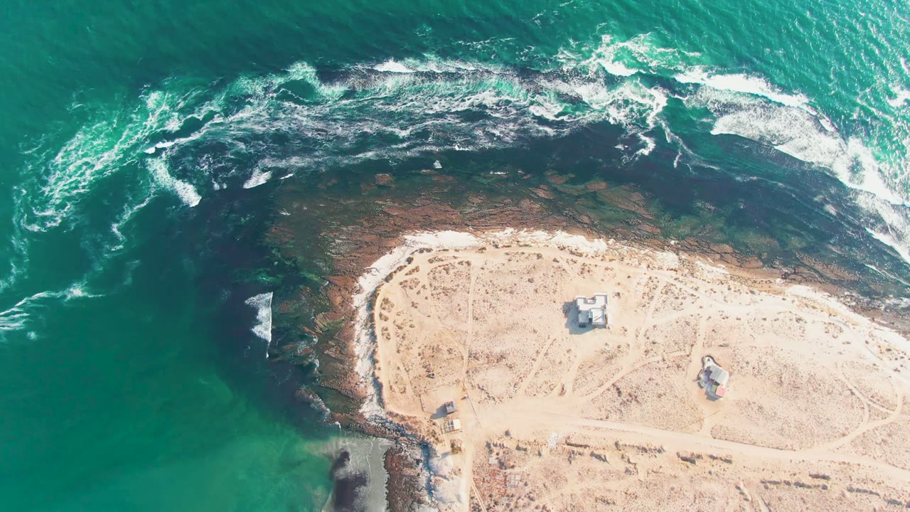 At 0:02, the La Bocana shoreline remains in a high overhead composition as waves break across the dark reef area near the small structures and empty sand.