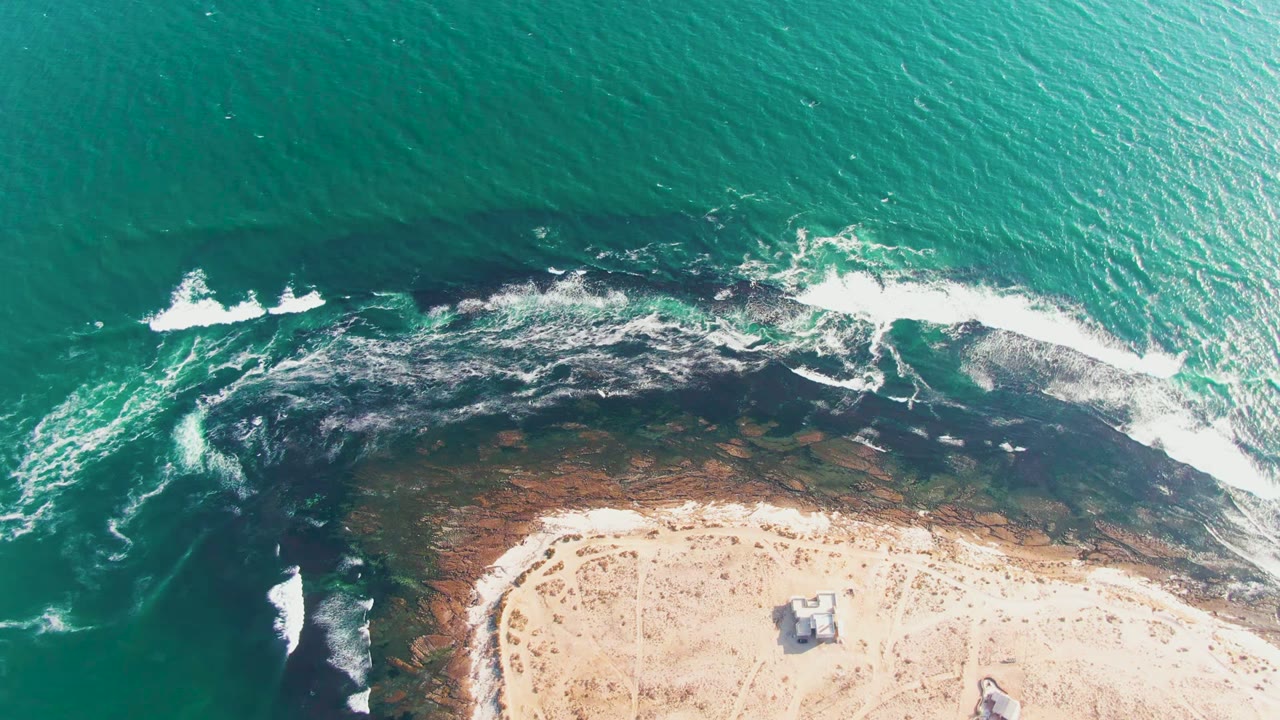 Overhead view of La Bocana turquoise water pushing white foam across dark rocky shoreline beside pale sand, with no visible people.