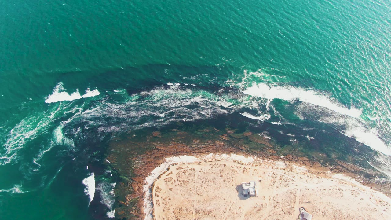 White waves spread along a dark reef edge next to sandy beach.