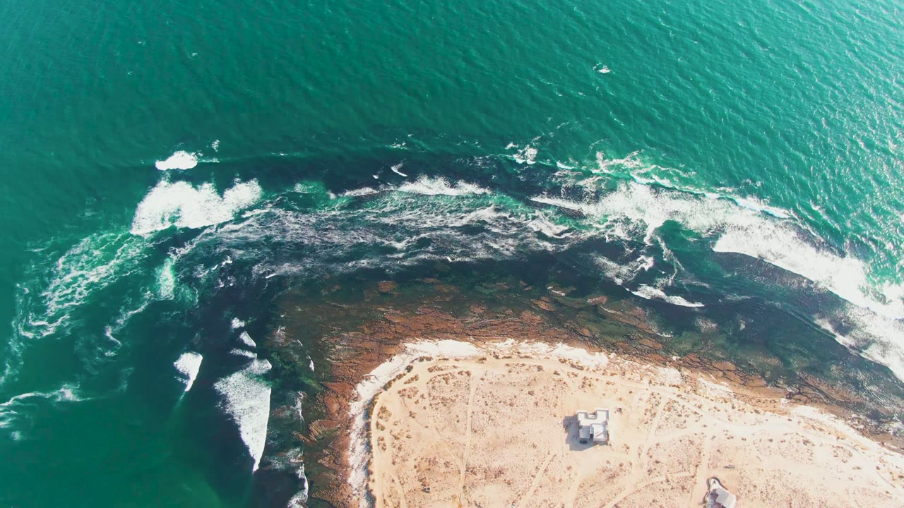 Overhead frame at La Bocana, Baja California Sur showing turquoise water, white surf, dark shoreline rocks, pale sand, and no visible people.