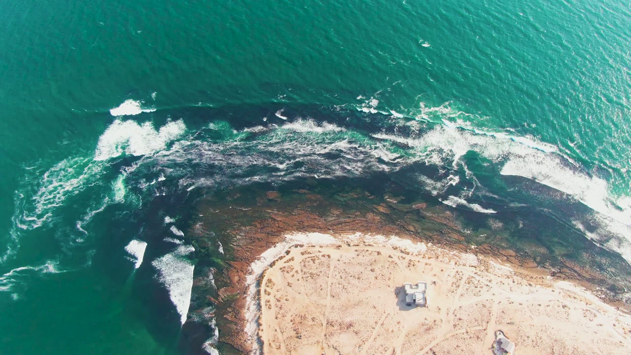 Aerial surf foam spreads across dark rocks next to a sandy beach
