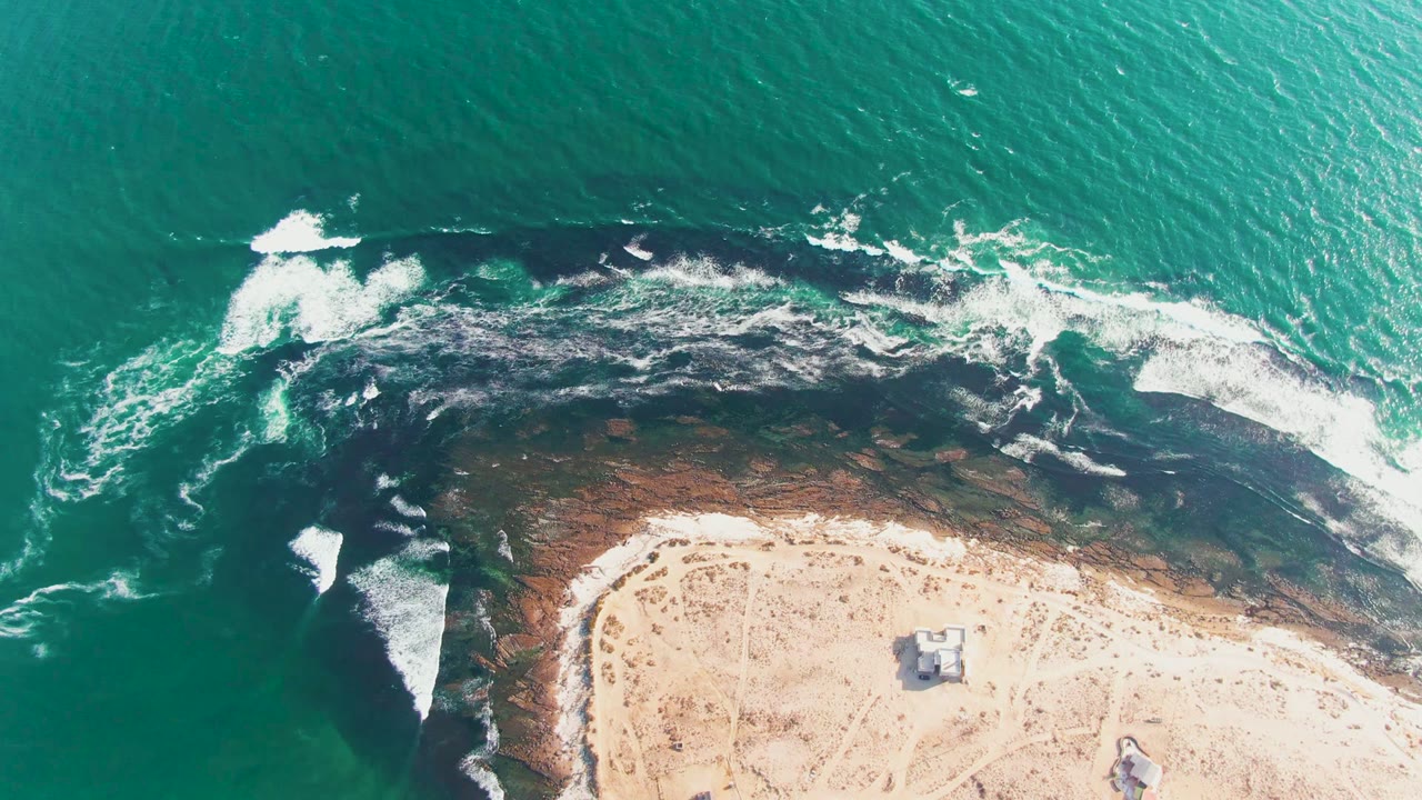 At 0:02, the overhead view of La Bocana shows a long white breaker crossing the dark rocks with green-blue water above and sandy beach below, with no visible people.