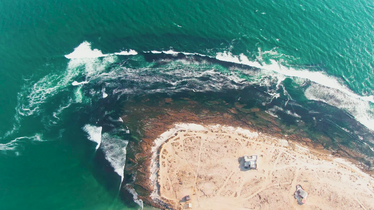 Aerial frame over La Bocana, Baja California Sur, with green ocean, white surf curling over dark rocks, pale sand below, small beach structures, and no visible people.