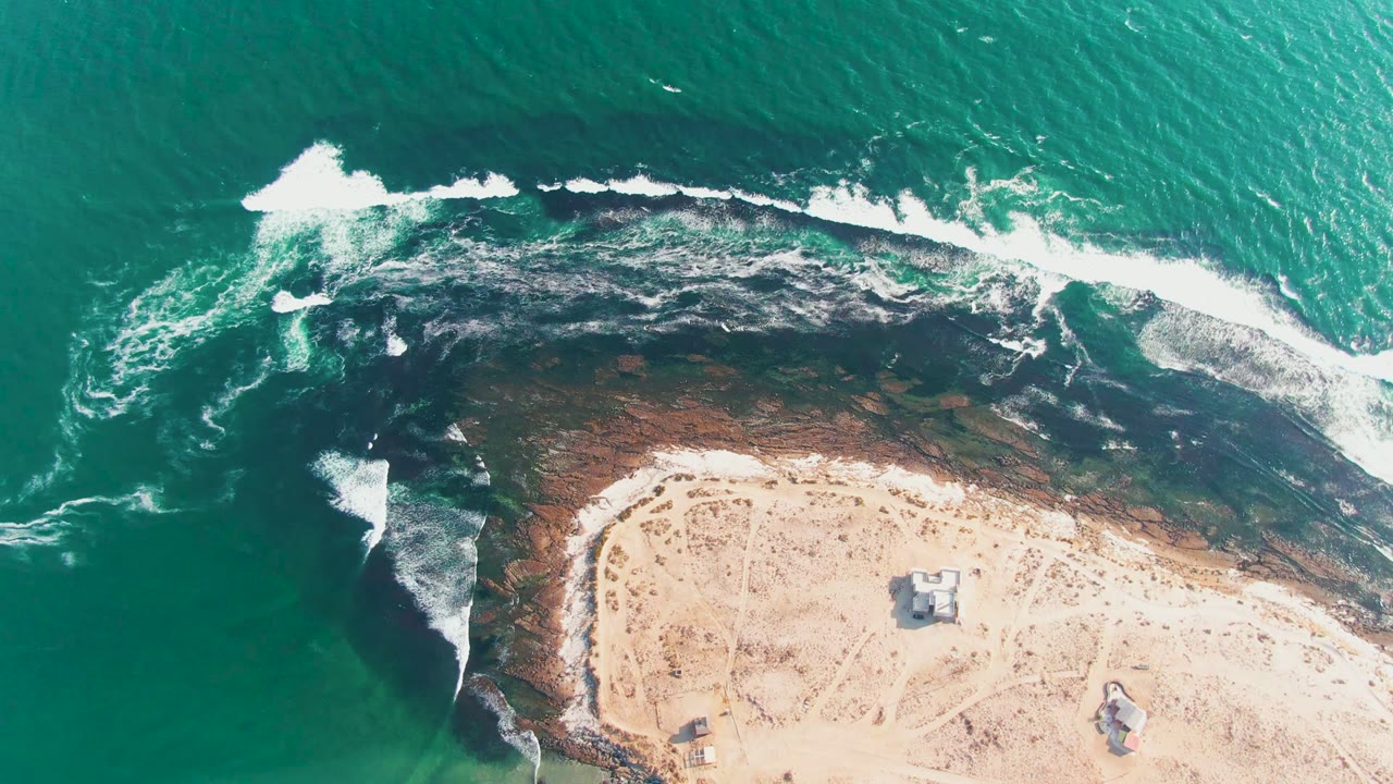 White wave foam spreads along a dark rocky shoreline next to sand.