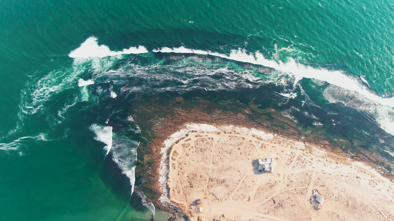 Aerial frame over the La Bocana coast with the breaking wave shifted farther along the rocky band, teal water above, pale beach below, small structures inland, and no visible people.