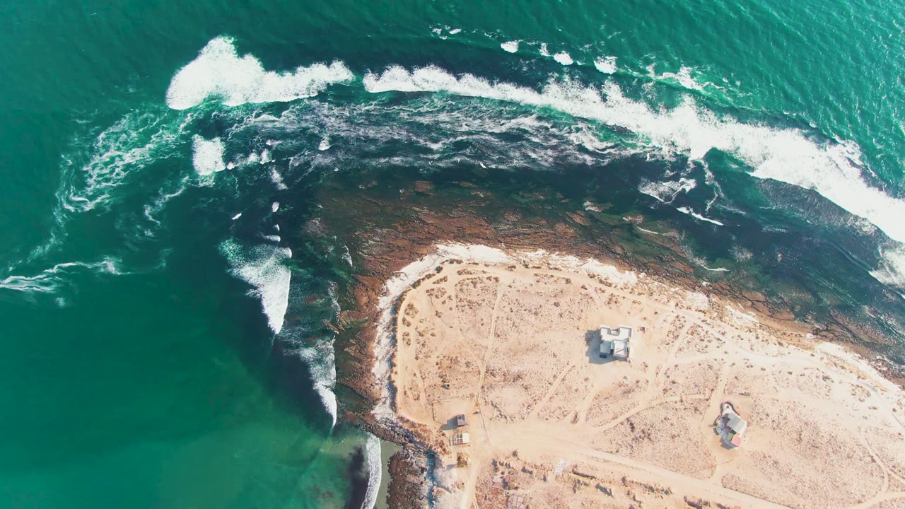 Aerial view over La Bocana, Baja California Sur, with turquoise ocean at left, white surf over dark rocky shallows, pale sand at lower right, and no visible people.