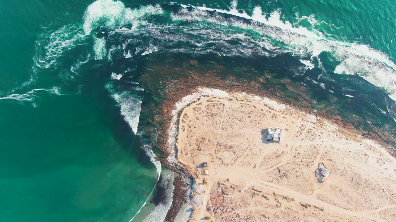 Aerial view over La Bocana, Baja California Sur, Mexico, with turquoise water on the left, white surf curling over dark rocks, pale sand to the right, small huts, and no visible people.