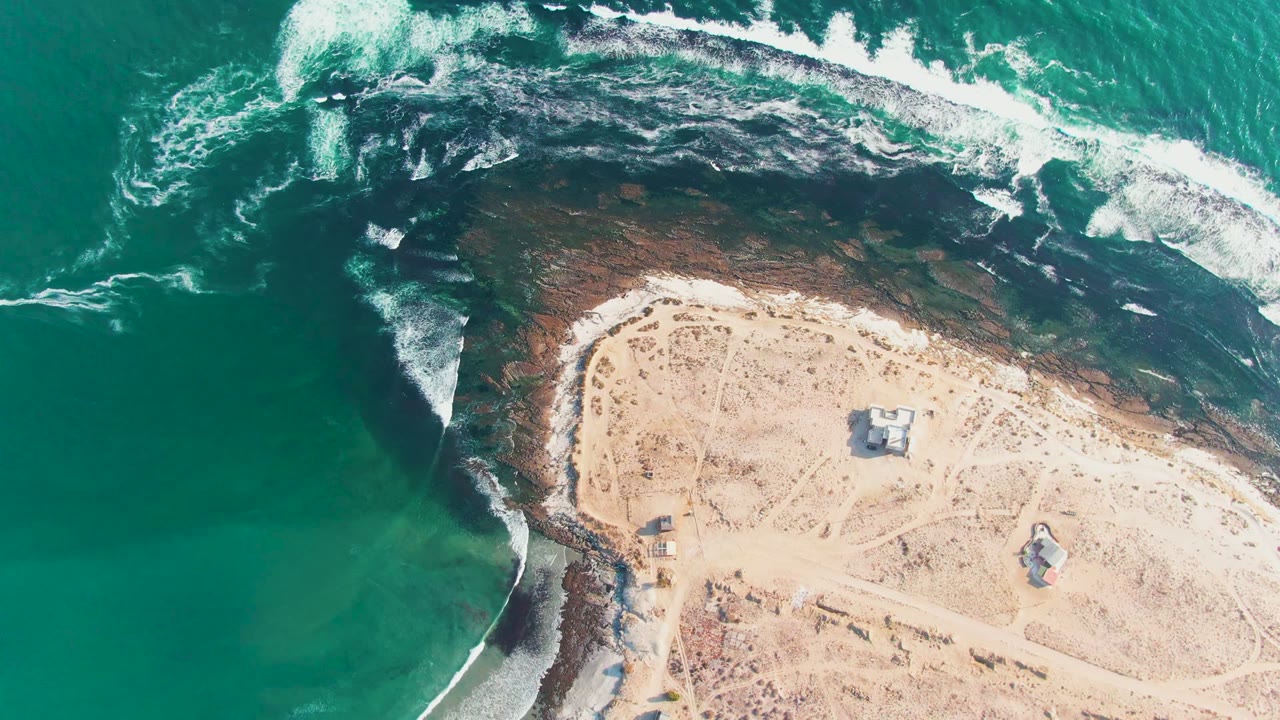 Top-down coastline with surf foam crossing dark rock and sand.