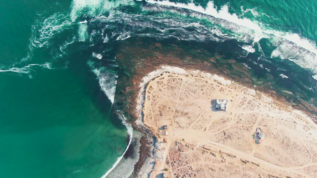 Drone view above La Bocana with foamy surf breaking along the top and left rock edge, a sandy point occupying the right half, several small huts or structures, and no visible people.