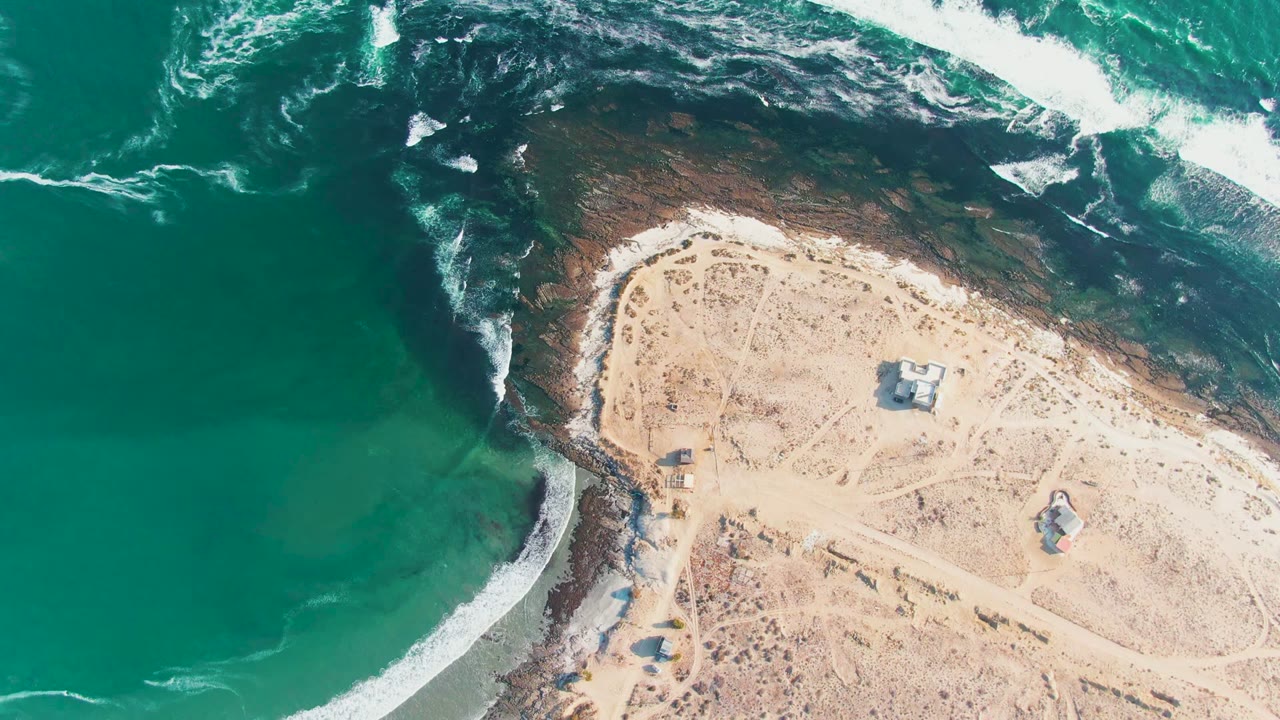 Aerial frame over La Bocana, Baja California Sur, with turquoise water at left, white surf on dark rocks, pale sand to the right, small structures inland, and no visible people.