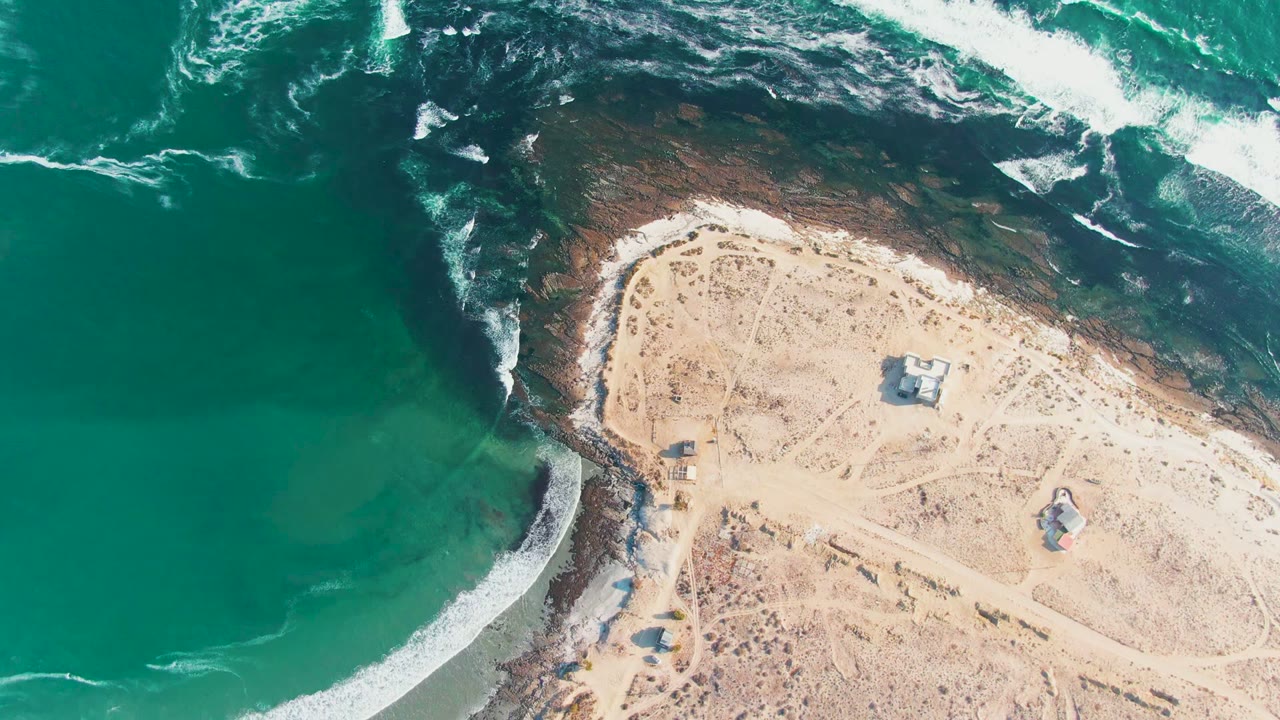 Drone view of waves curving around dark rocks beside sandy land.