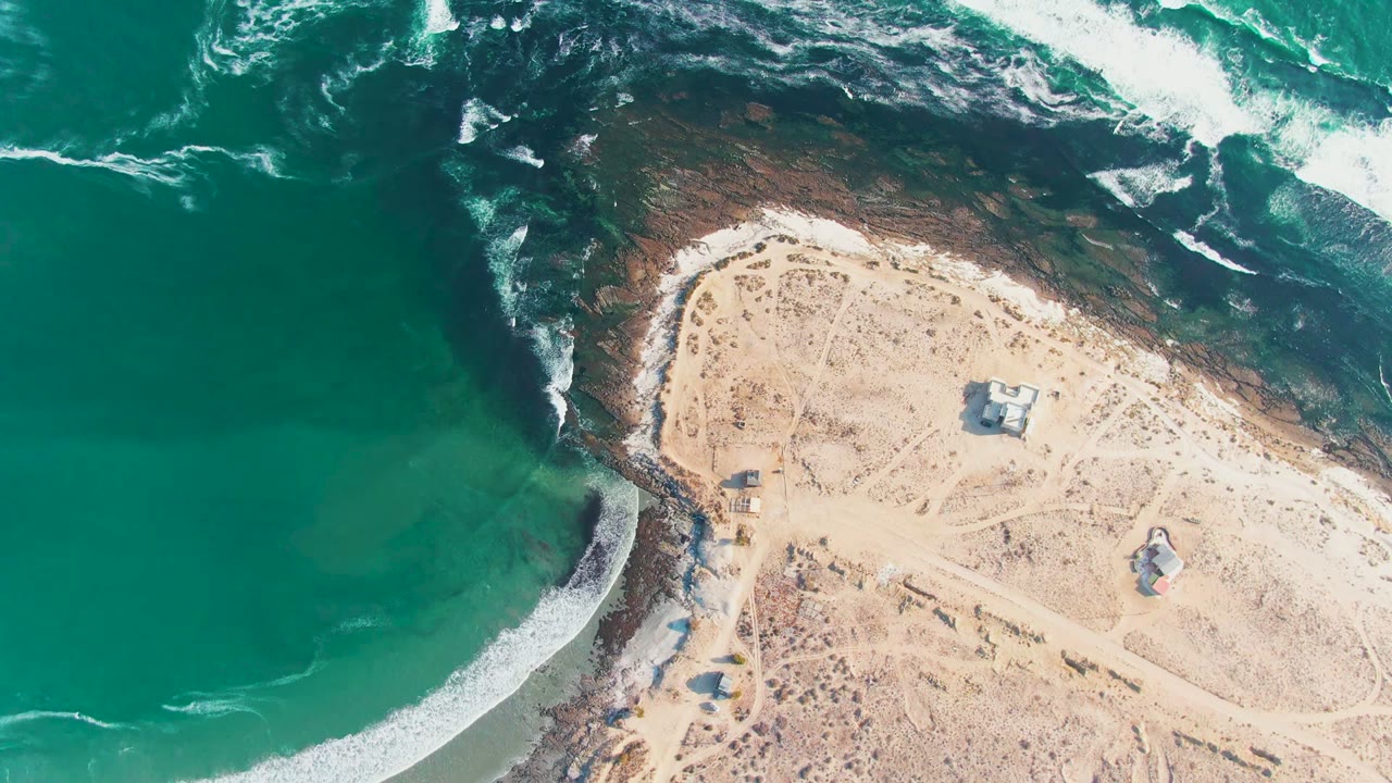 Top-down frame over La Bocana, Baja California Sur, with darker water near rocks at the top, a pale sandy point on the right, scattered low structures, dirt tracks, and no visible people.