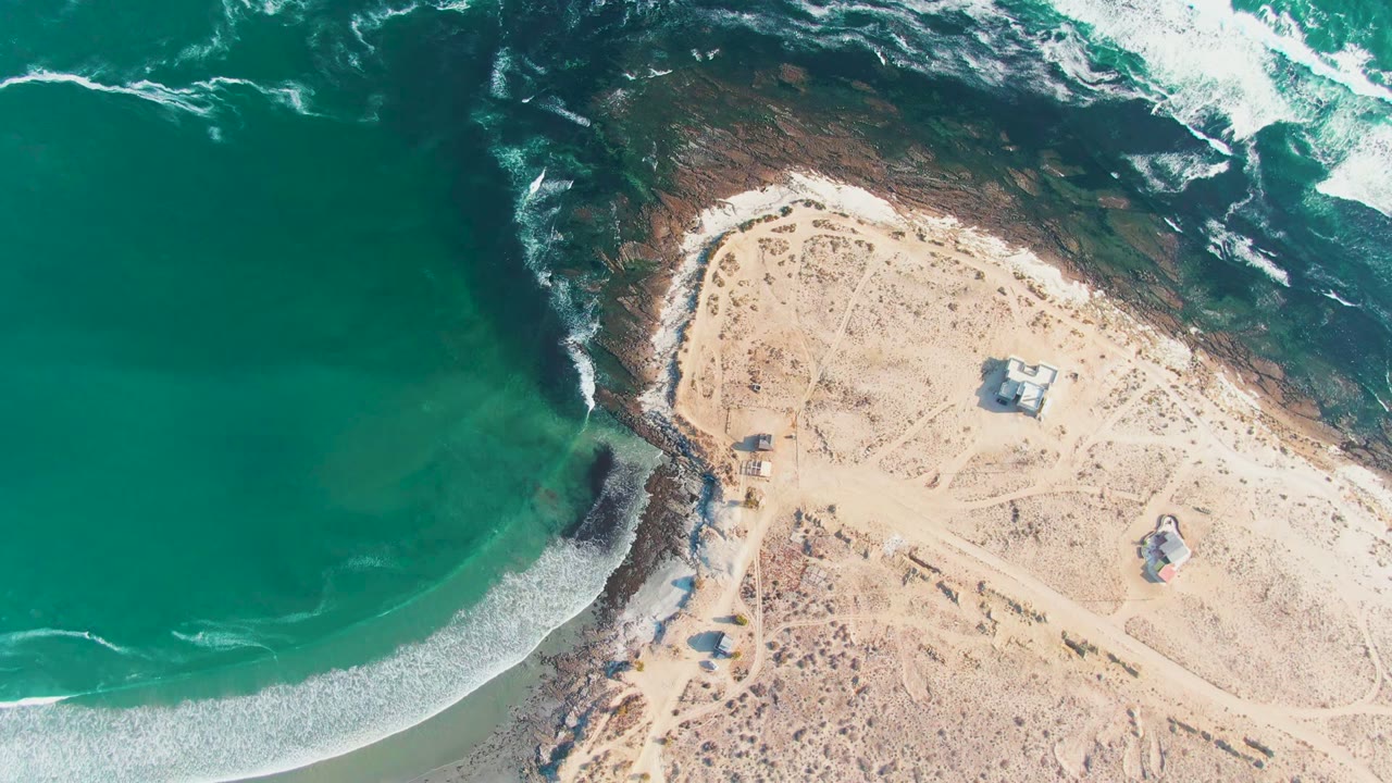 High aerial view over La Bocana, Baja California Sur, with green-blue ocean, white surf, a sandy rocky point, small structures inland, and no visible people.