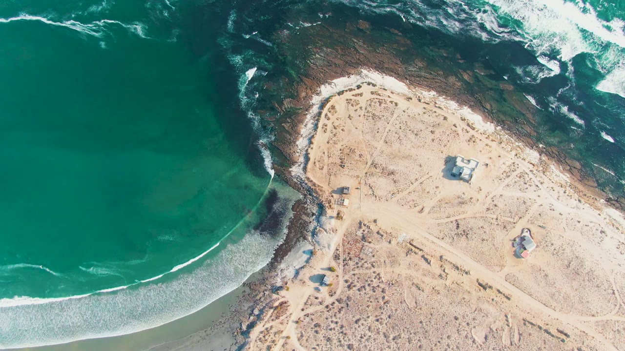 High overhead view at La Bocana with teal water on the left, dark rock shelves at the top, pale sand to the right, and no visible people.