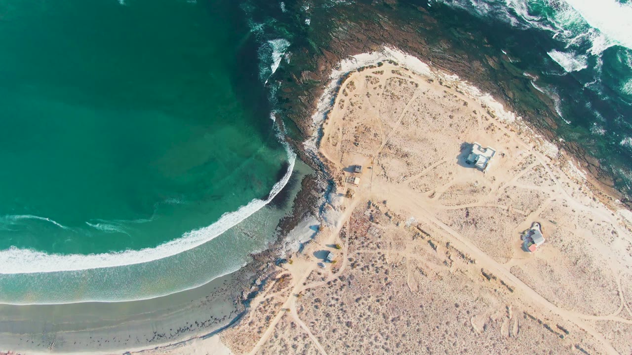 Overhead frame from La Bocana, Baja California Sur, showing green-blue water, a white foam line, pale sand, rocky coast, small structures, and no visible people.