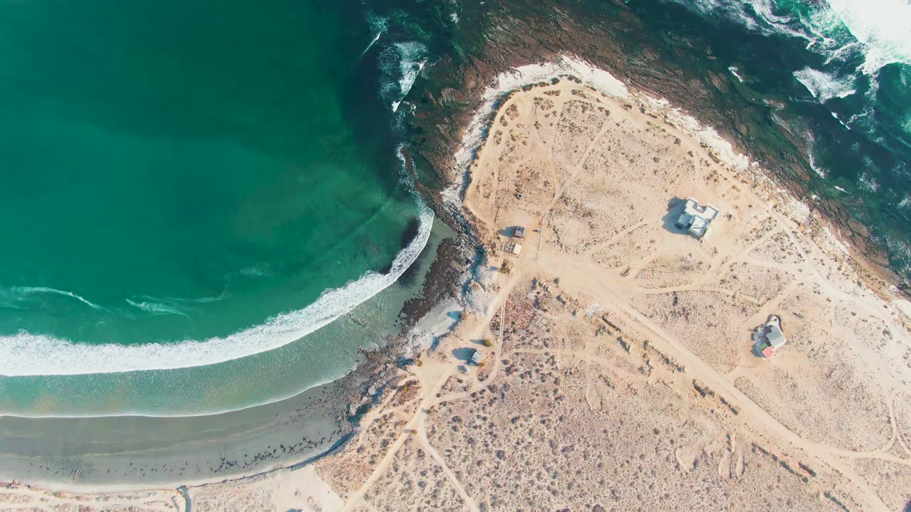 Top-down La Bocana frame showing a broad green water area, a clean white breaker line, dark rocky shoreline, pale sand, scattered small structures, and no visible people.
