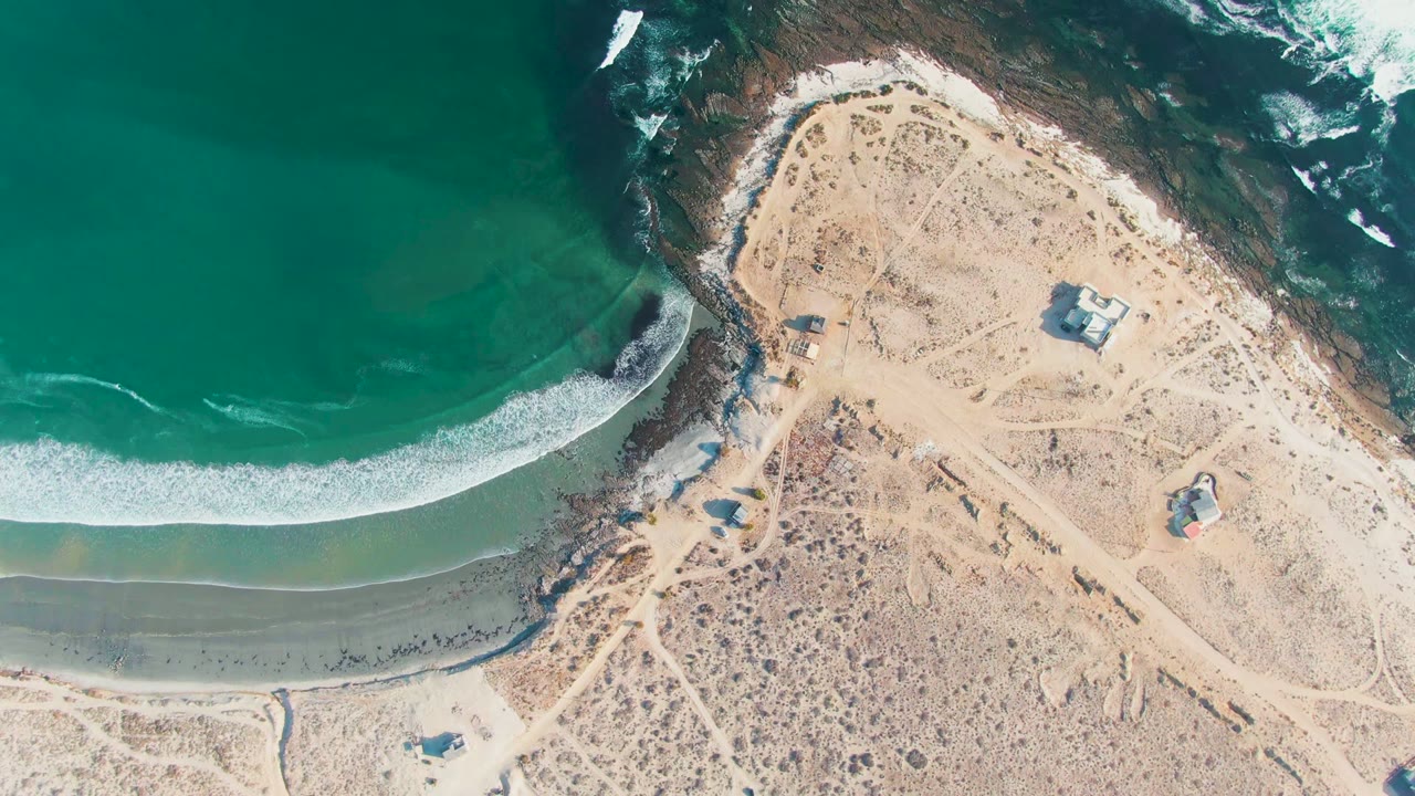 At 0:00, an overhead view of La Bocana shows turquoise water curling toward a sandy beach beside pale desert ground, with no visible people.