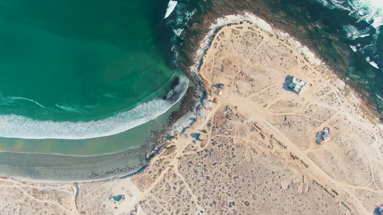 Aerial coastline with foam advancing across a narrow sandy shore near desert tracks.