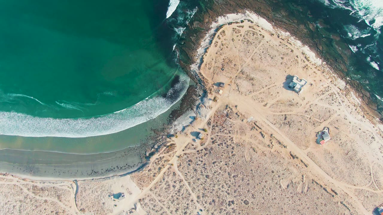 At 0:02, green-blue ocean water and a white breaker arc along the La Bocana beach beside open desert terrain and scattered small structures, with no visible people.