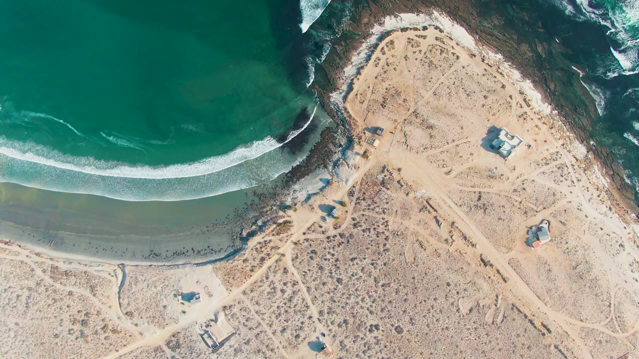 Overhead frame of La Bocana, Baja California Sur, showing green-blue water, a white surf line, pale sand, dirt tracks, rocky edges, small structures, and no visible people.
