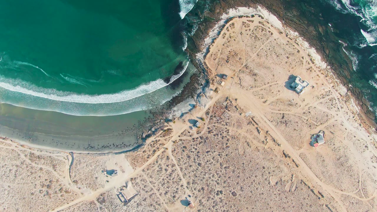 Aerial coastline with a curved wave band, wet sand, desert tracks, and small buildings.