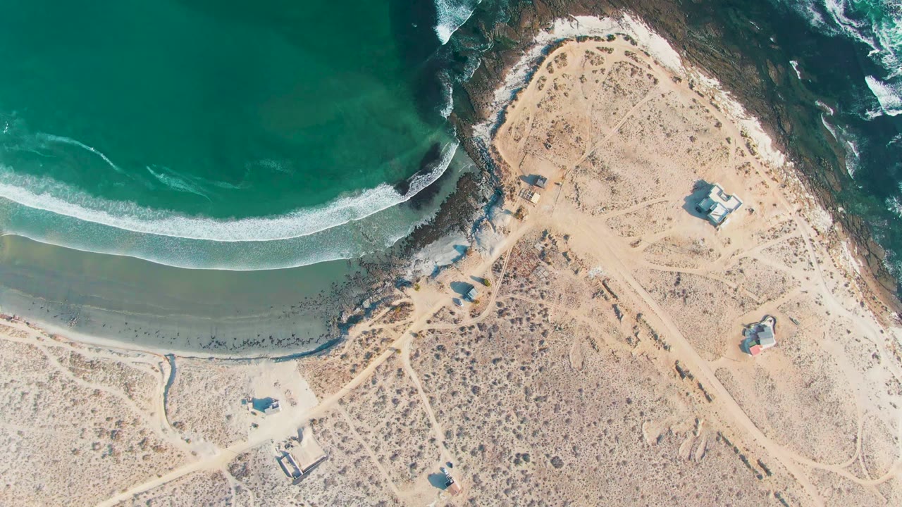 At 0:02, the overhead La Bocana frame shows the ocean filling the top, a clean arc of white foam at the beach, dry sandy land with tracks below, small structures, and no visible people.