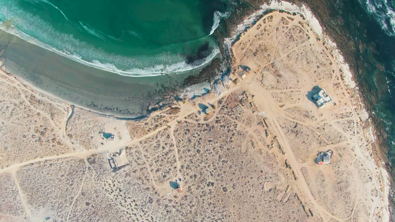 Overhead frame from La Bocana, Baja California Sur, showing teal water at the top, a pale sandy shoreline, arid scrubland crossed by dirt paths, scattered small structures, and no visible people.
