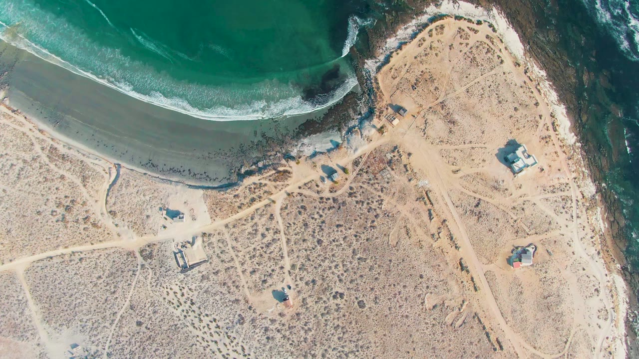 Aerial shoreline view with blue-green surf, pale sand, dirt tracks, and isolated coastal buildings.