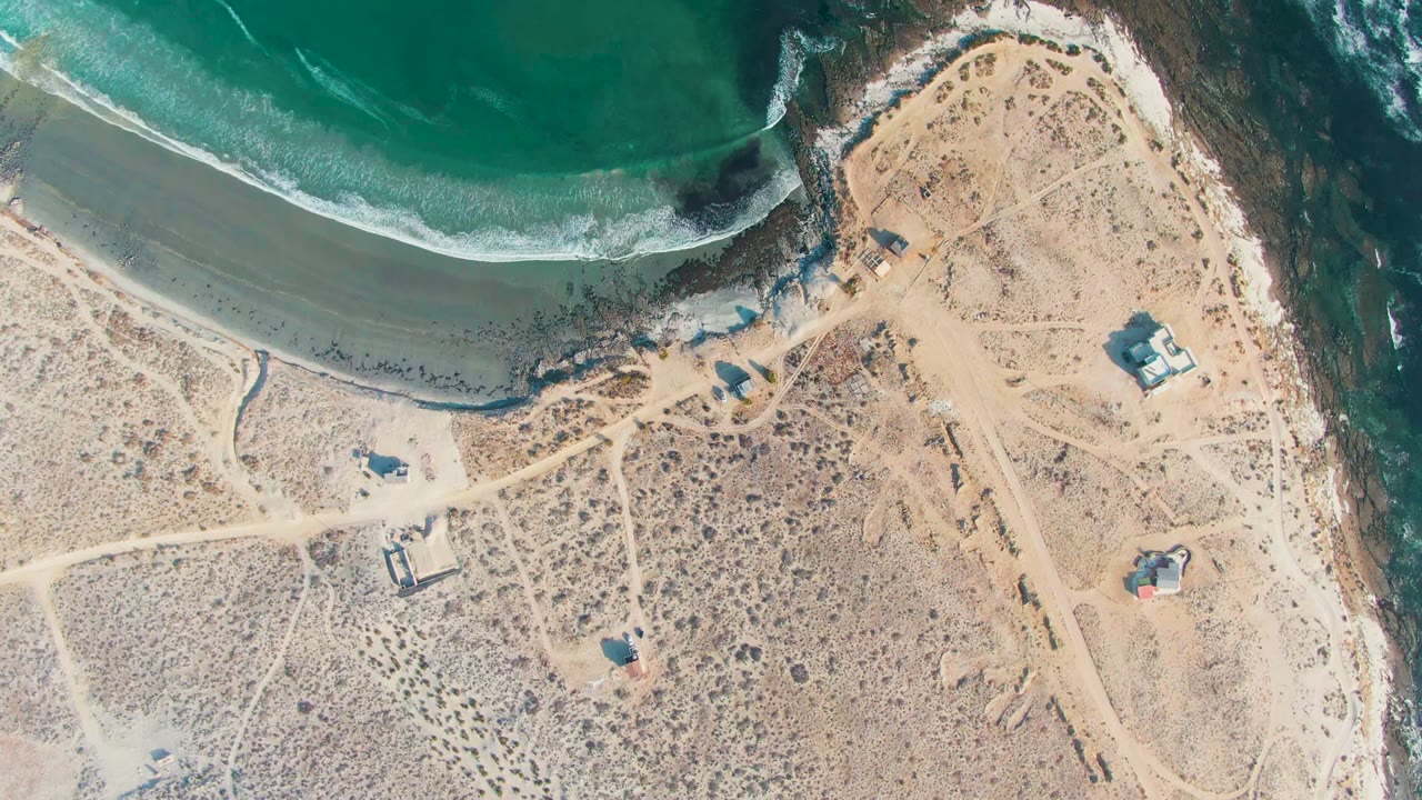 At 0:02 in La Bocana, the overhead composition shows turquoise water pressing toward a broad sand strip, darker rock along the right shoreline, sparse scrub, pale paths, and no visible people.