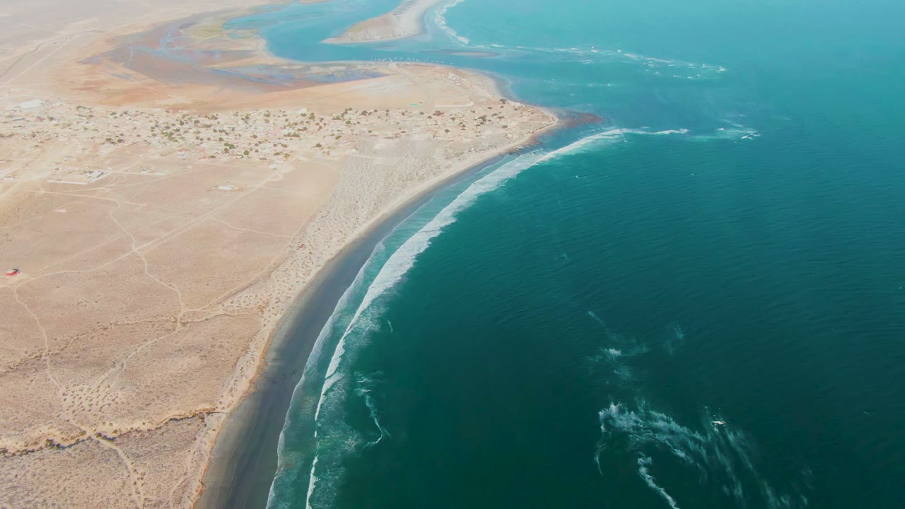 Aerial frame over La Bocana, Baja California Sur showing tan desert shore curving beside blue-green ocean with white surf lines and no visible people.