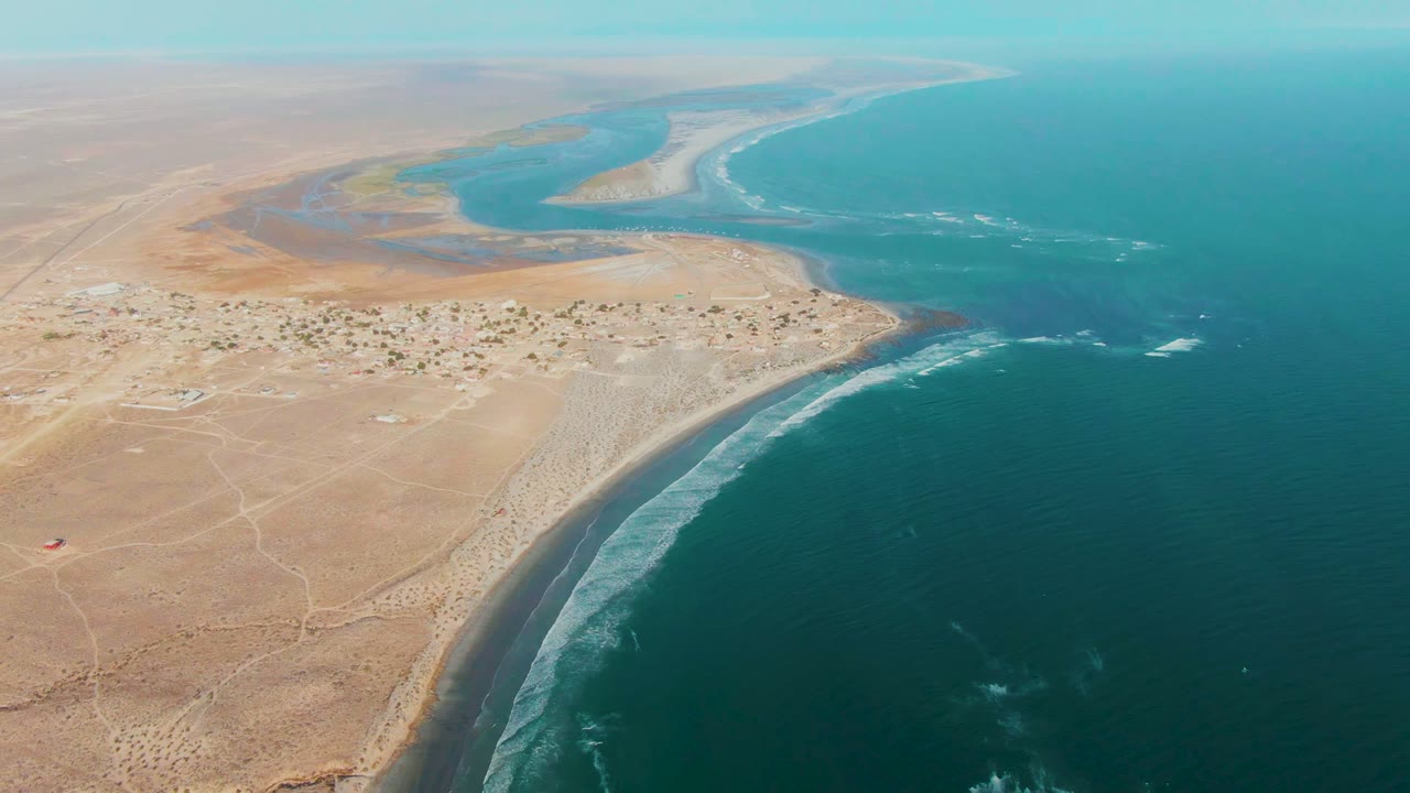 Wide aerial view over La Bocana, Baja California Sur, with tan desert in the foreground, dark blue ocean at right, a river estuary beyond the settlement, and no visible people.