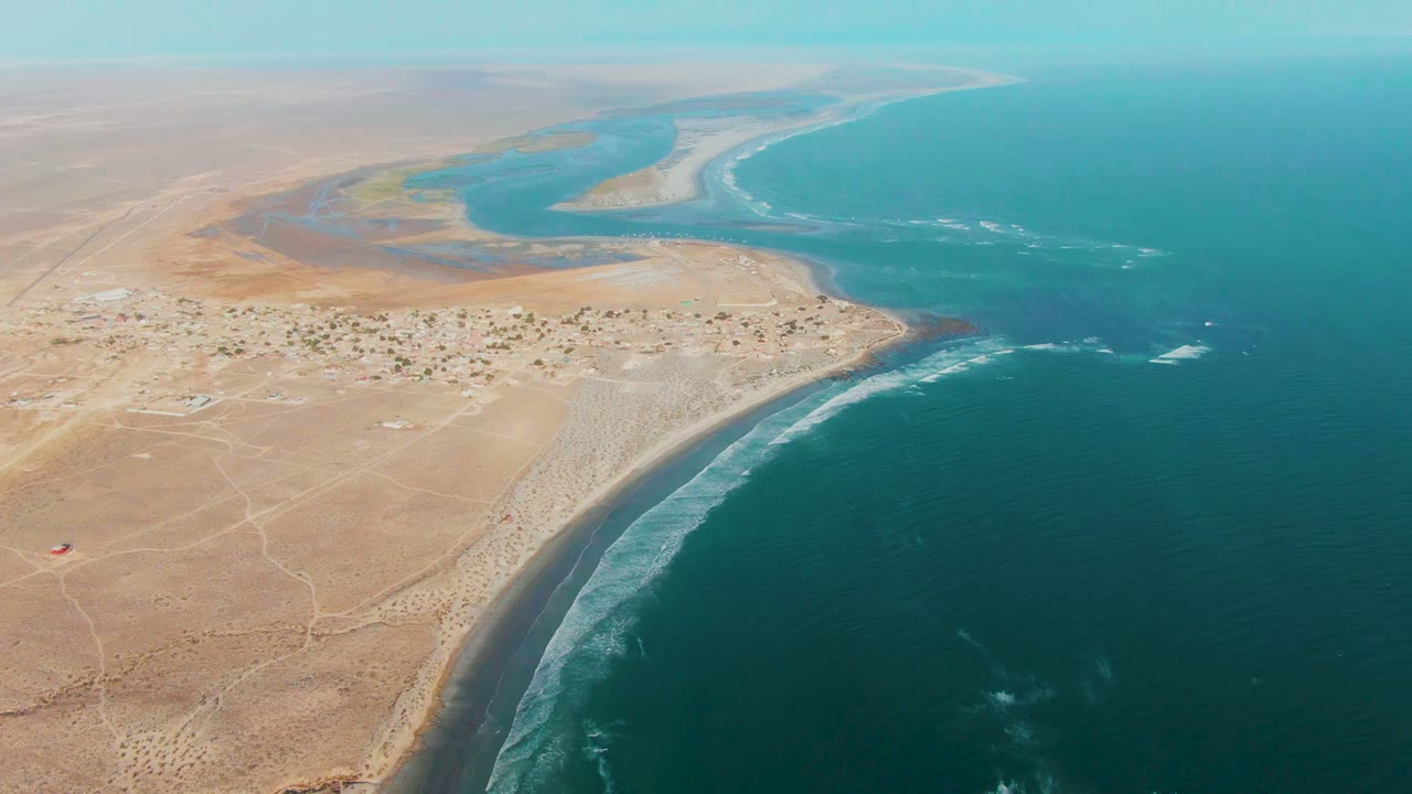 Aerial coastline view with curved surf, arid land, settlement, and blue-green estuary.