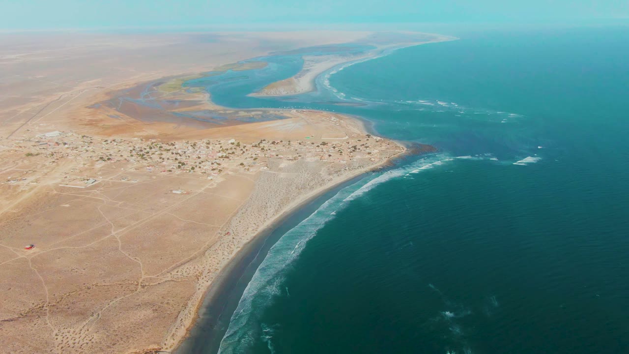 High aerial view of La Bocana with ocean waves tracing the shore, pale desert flats and a small settlement on the left, blue estuary water beyond, and no visible people.