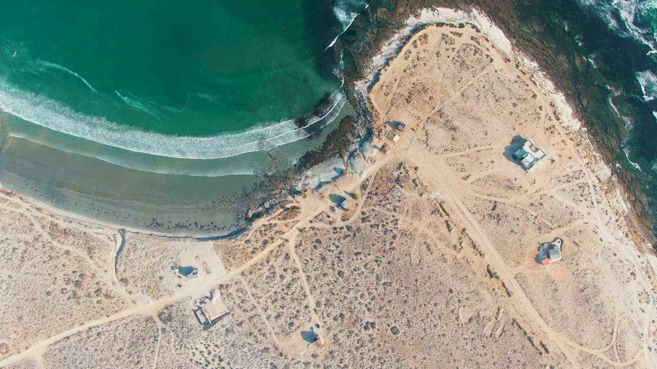 Opening aerial frame over La Bocana, Baja California Sur, showing turquoise water, a curved surf line, sandy beach, dry scrubland, dirt tracks, small structures, and no visible people.