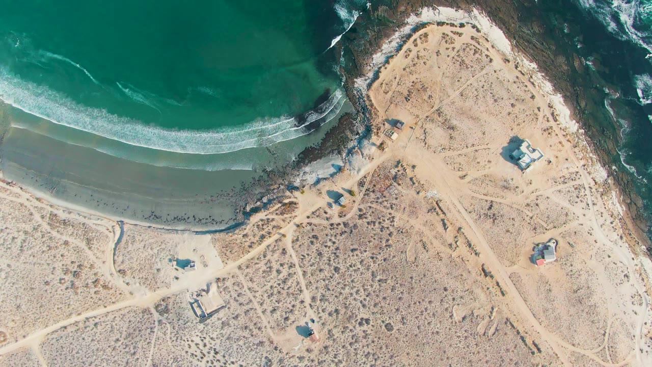 Aerial shoreline with white wave band, sandy beach, dirt roads, and huts