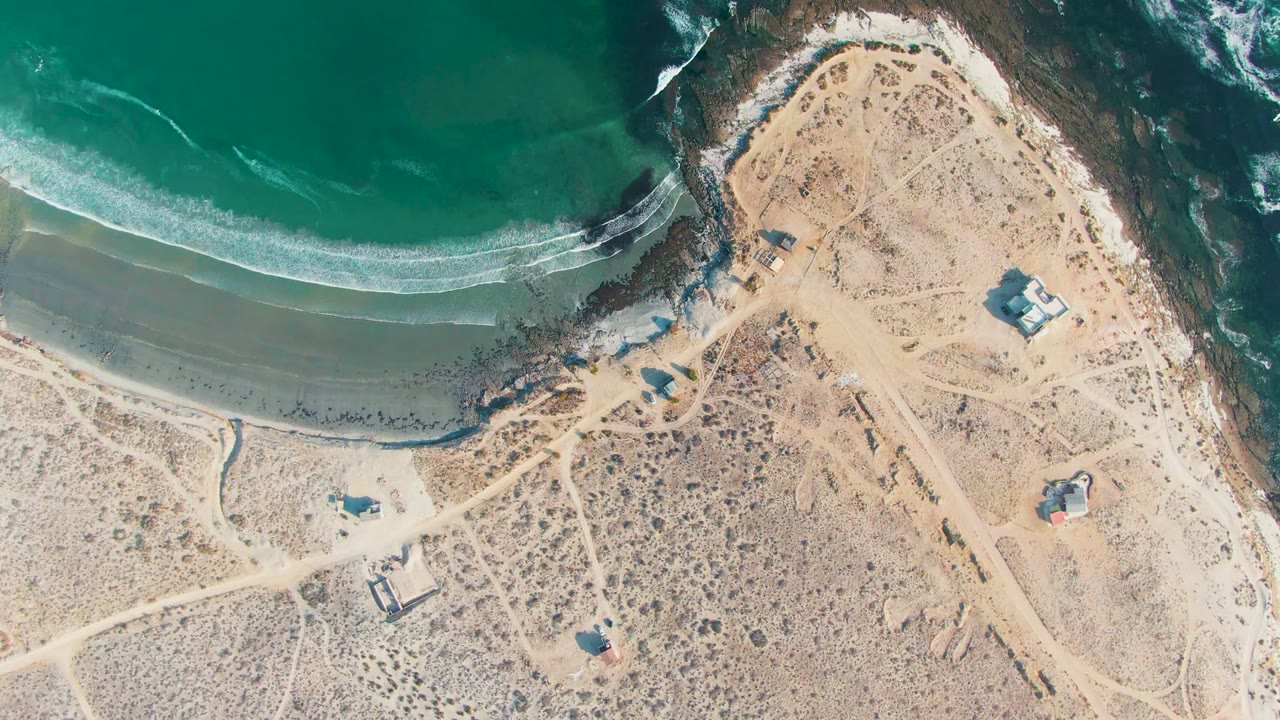 At 0:02, the La Bocana coast remains in a top-down frame with turquoise water, a thin foam line, pale sand, scrub-dotted land, small buildings, tracks, and no visible people.