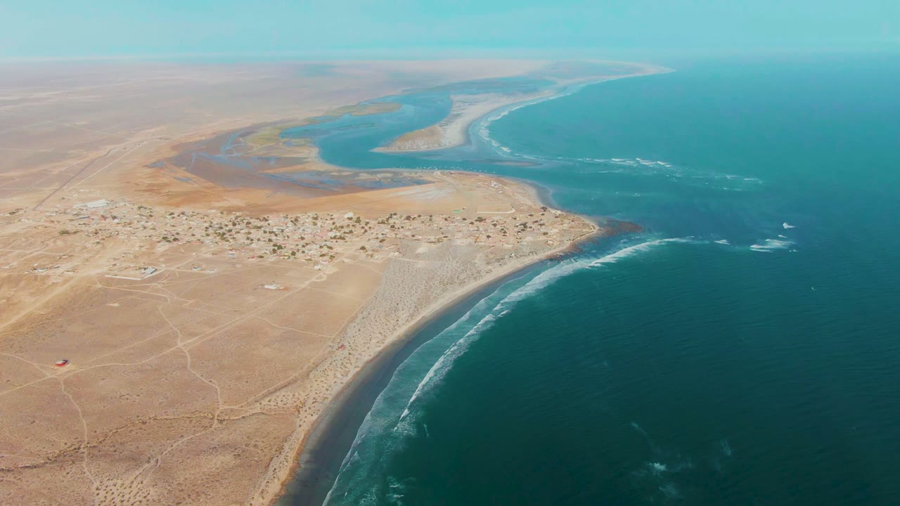Opening aerial frame over La Bocana, Baja California Sur, with tan desert foreground, a small shore settlement, lagoon water, ocean surf, and no visible people.