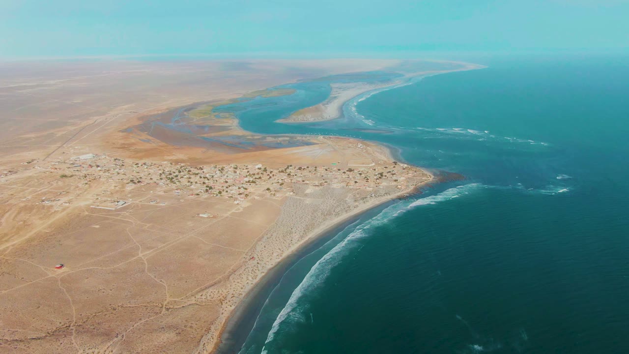 Aerial frame over La Bocana, Baja California Sur showing tan desert, a small shore village, curved beach, lagoon, ocean waves, a small red vehicle on open sand, and no visible people.