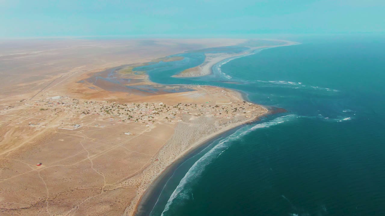 Aerial frame over La Bocana showing more open ocean to the right, the village and pale beach below, lagoon shallows beyond the settlement, and no visible people.