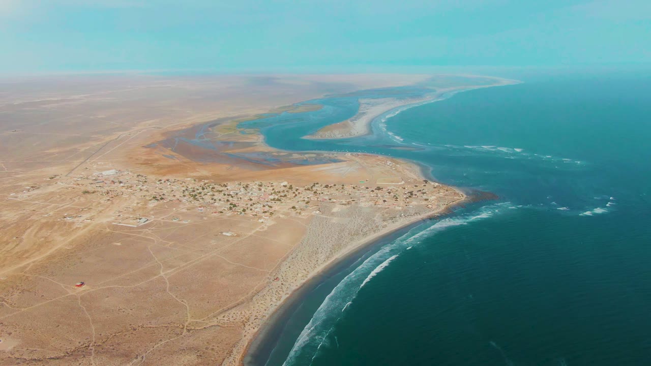 Aerial view over La Bocana, Baja California Sur, with tan desert terrain, a small coastal settlement, surf along the beach, blue ocean, inland water channels, and no visible people.
