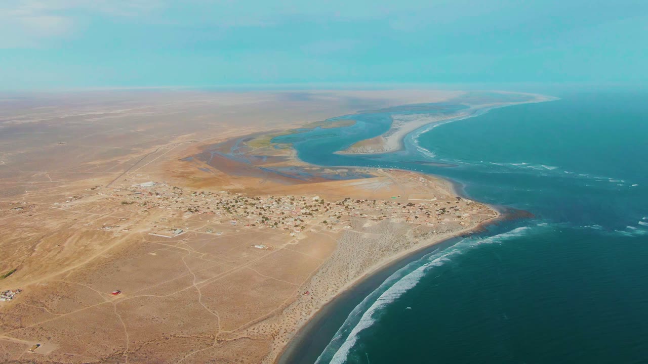High aerial view over La Bocana, Baja California Sur, with sandy desert in front, a small settlement near the coast, an estuary curving into blue ocean, and no visible people.