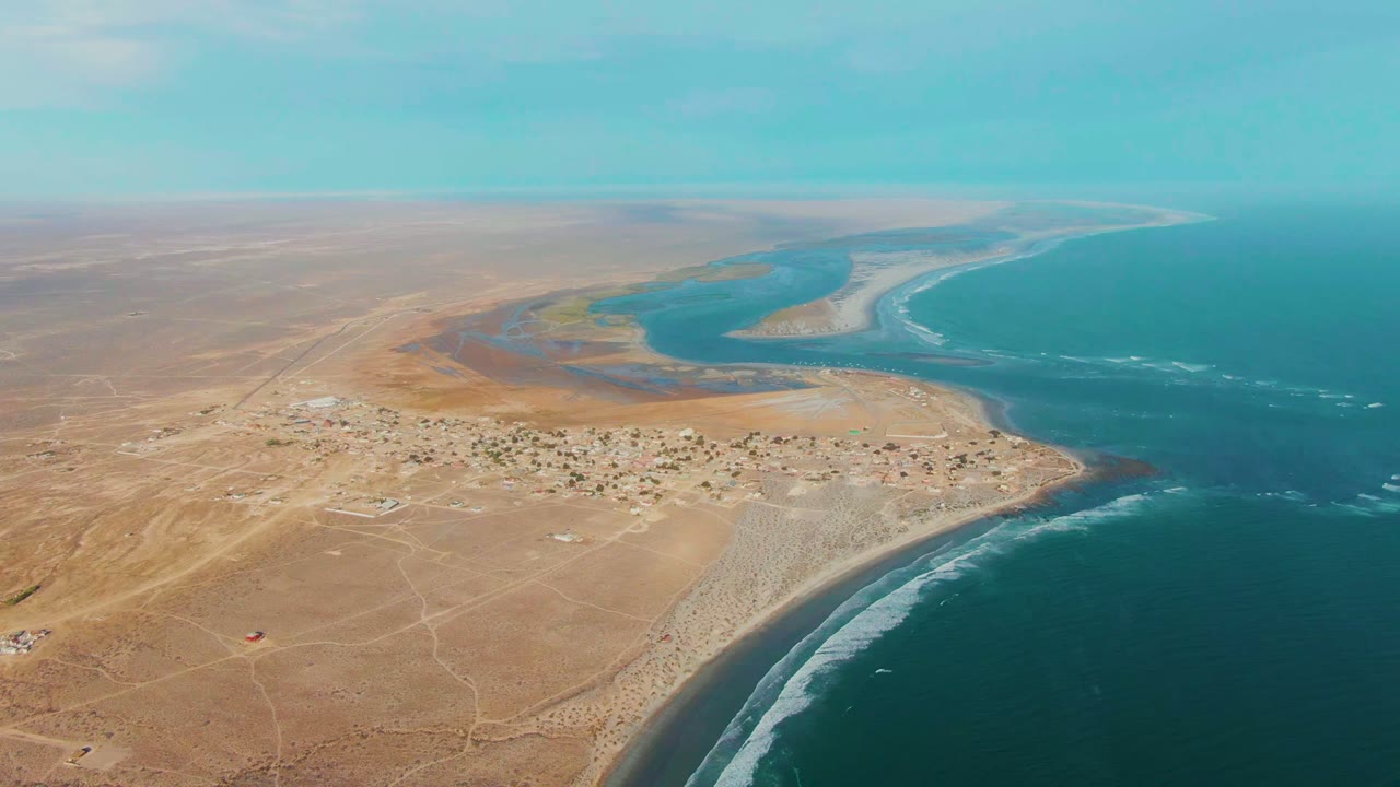 Drone view of sandy desert, coastal homes, estuary, and breaking waves.