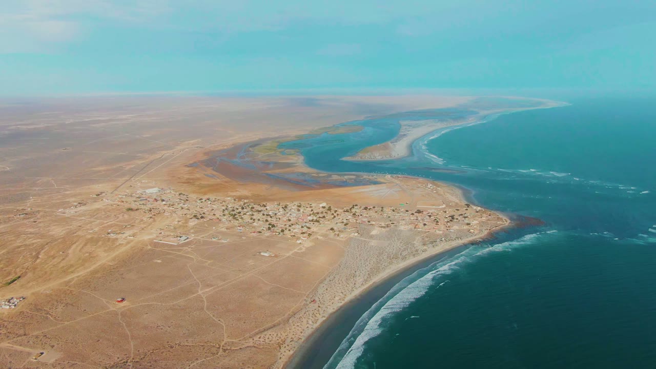 Wide drone view of La Bocana with the settlement slightly lower in frame, tan desert stretching left, turquoise water beyond the estuary, white wave bands, and no visible people.