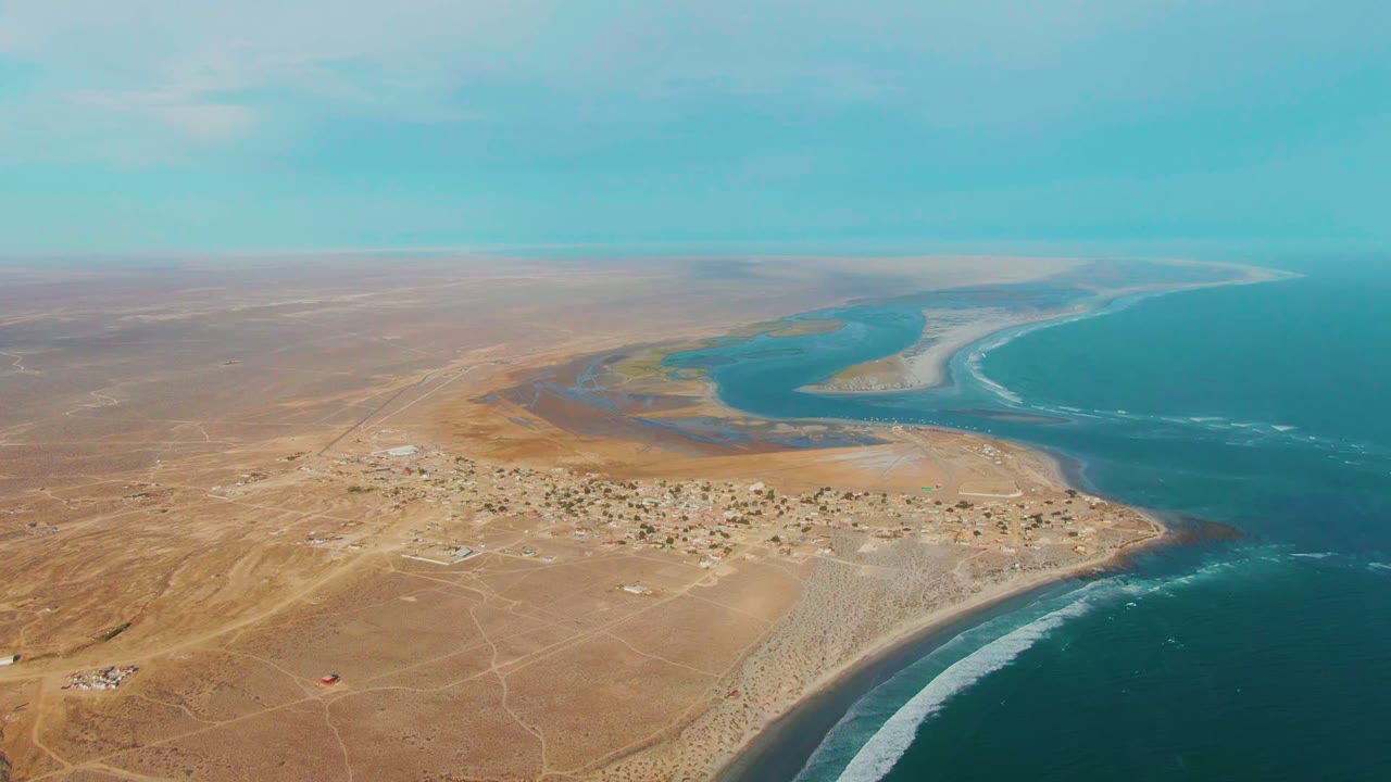 Opening aerial frame over La Bocana, Baja California Sur, showing sandy desert foreground, a small coastal settlement, blue ocean, white surf, and no visible people.