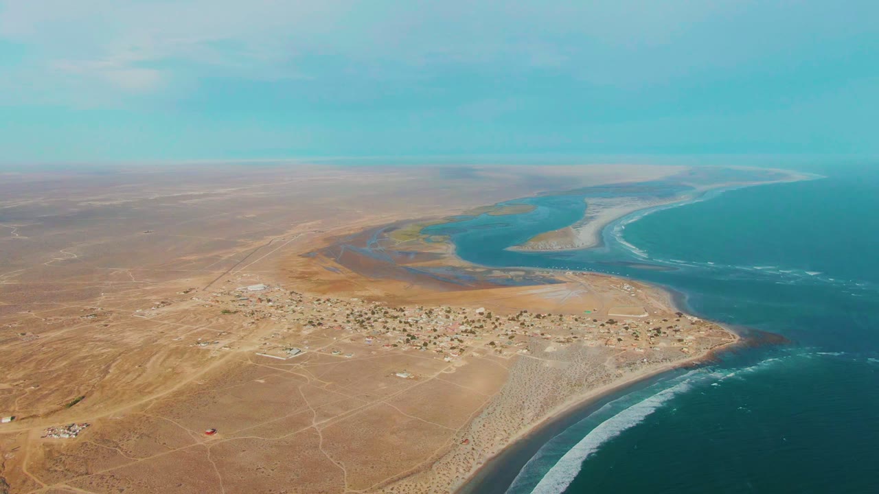 Wide aerial coastline with desert village, surf, and lagoon water beyond