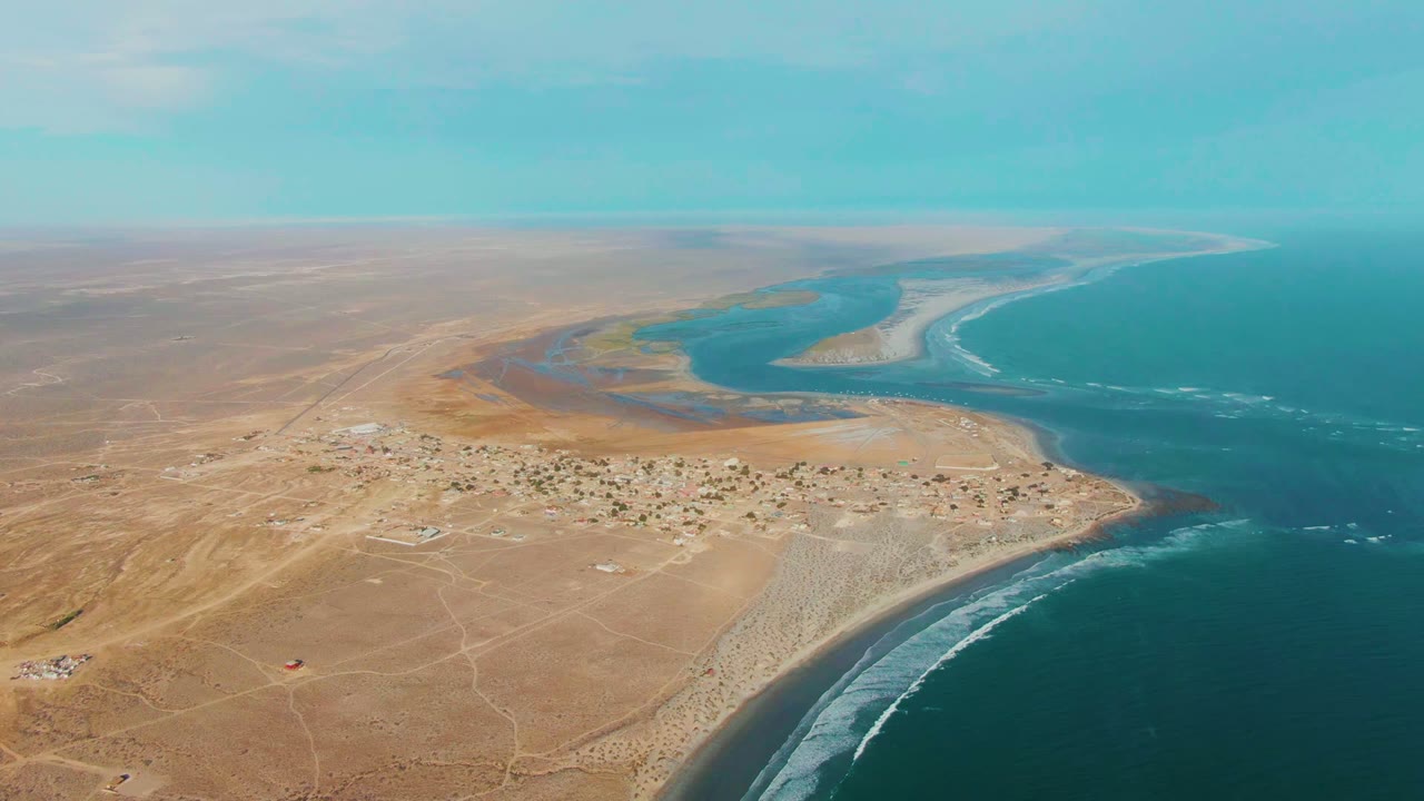 Opening aerial frame over La Bocana, Baja California Sur, with tan desert roads, a small settlement, blue lagoon water, surf lines, and no visible people.