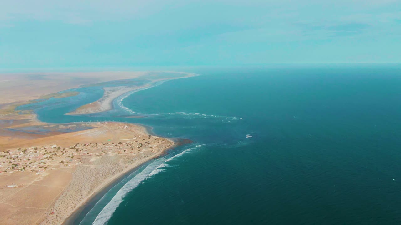 Aerial frame from La Bocana showing the river mouth and sandy spit stretching into the distance, clustered low buildings near shore, dark blue ocean, and no visible people.